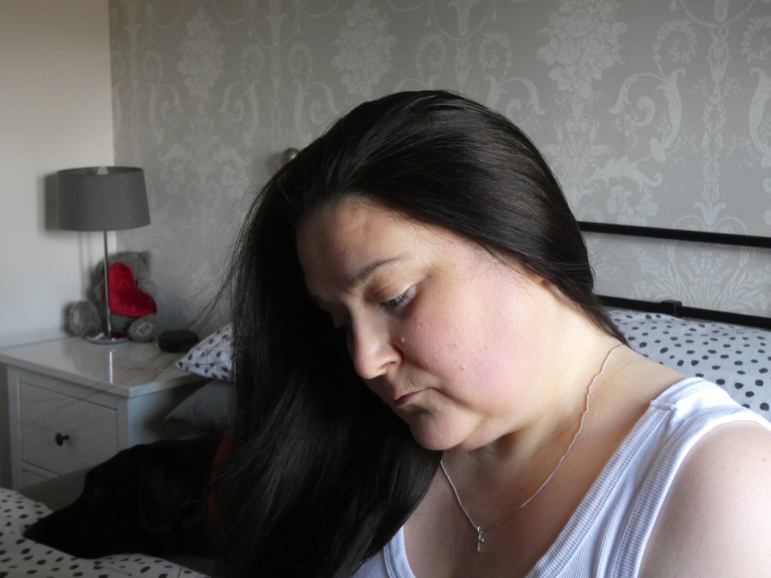 Lady sat on bed wearing white vest top around her neck is a necklace with a silver cross the lady has dark brown hair and the photograph is taken side on giving a side profile